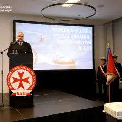 Mężczyzna przemawia przy mównicy z logo SAR. Na ekranie morska służba poszukiwania i ratownictwa.