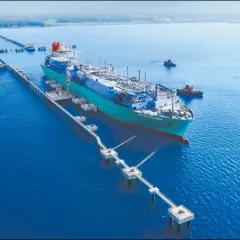 Pływający terminal LNG przycumowany do nabrzeża, z holownikami na wodzie.