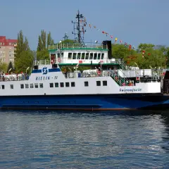 Prom "Bielik II" z pasażerami na pokładzie, płynący po wodzie, z budynkami w tle.