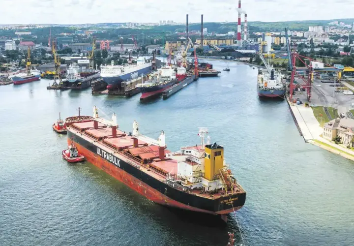 Duży statek towarowy "ULTRABULK" manewruje w porcie z pomocą holowników, w tle stocznie i miasto.