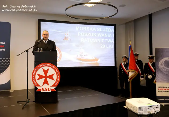 Mężczyzna przemawia przy mównicy z logo SAR. Na ekranie morska służba poszukiwania i ratownictwa.