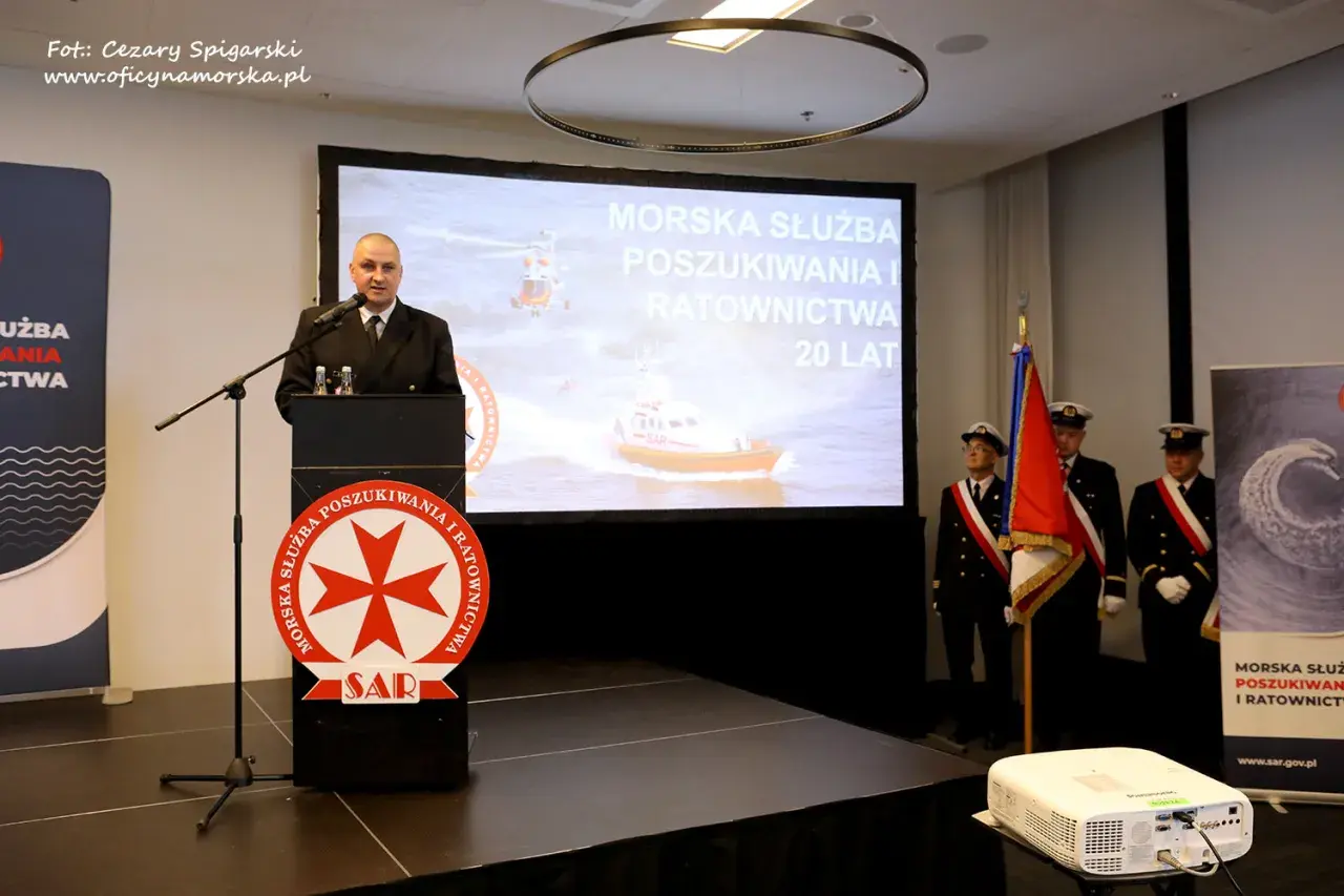 Mężczyzna przemawia przy mównicy z logo SAR. Na ekranie morska służba poszukiwania i ratownictwa.